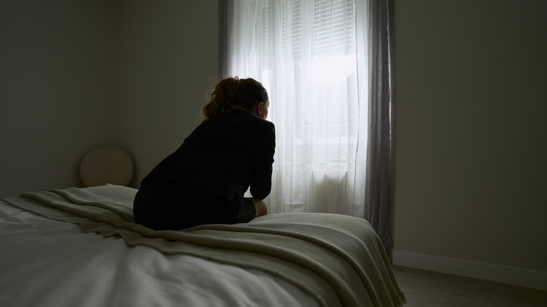 Woman facing window with curtains sitting on the edge of a bed.