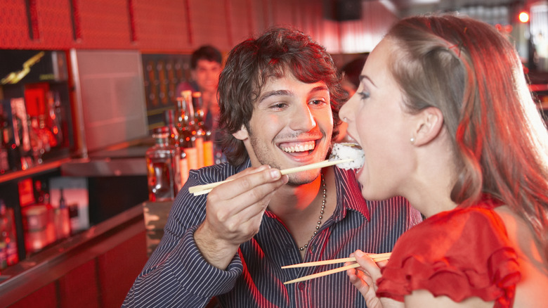 A man feeding sushi to a woman with chopsticks in a Japanese restaurant.