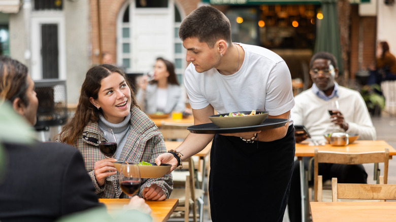 A waiter brings plates of food to a female diner sitting outside a restaurant.