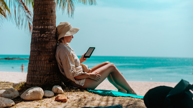 A woman with an e-reader sitting under a palm tree on the beach.