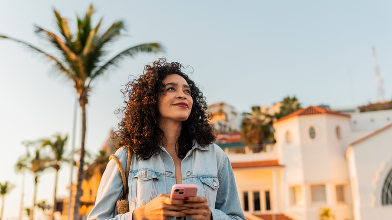A young woman holding a mobile phone in a beach town in front of white buildings and a palm tree.