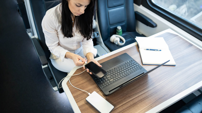 A woman charging her phone using a power bank while seated at a table on a train