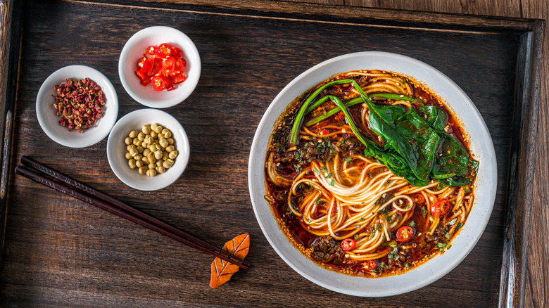 Bowls of noodles and other ingredients stand on a table with chopsticks
