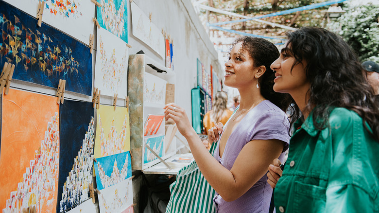 Two woman smiling at paintings at an outdoor market stall.