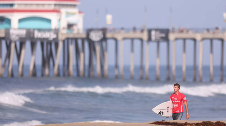 Surfer in red carrying a board with blurry waves and pier in the background