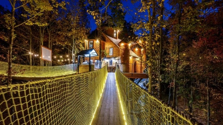 Lit suspension bridge leading to a wooden house surrounded by trees
