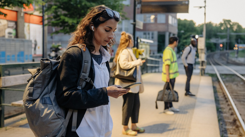 Woman looking down at phone wearing AirPods while standing among people waiting for train at station