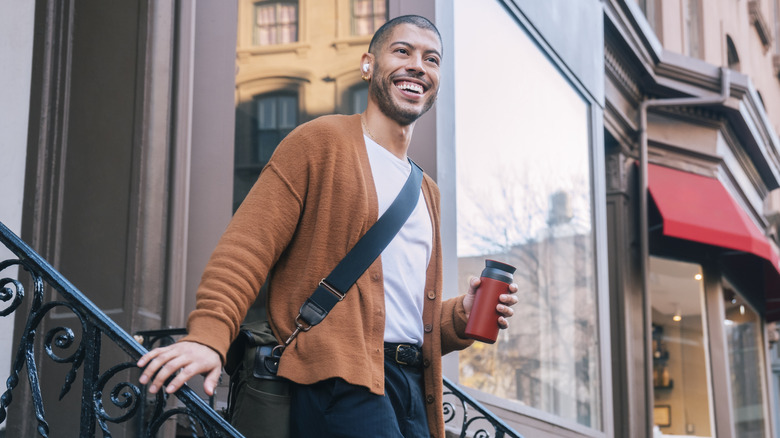 Man wearing AirPods smiling as he holds coffee and walks down the steps of a brownstone building