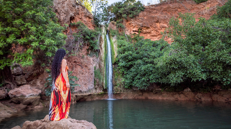 A woman in a long multi colored dress standing on a rock looking at a waterfall