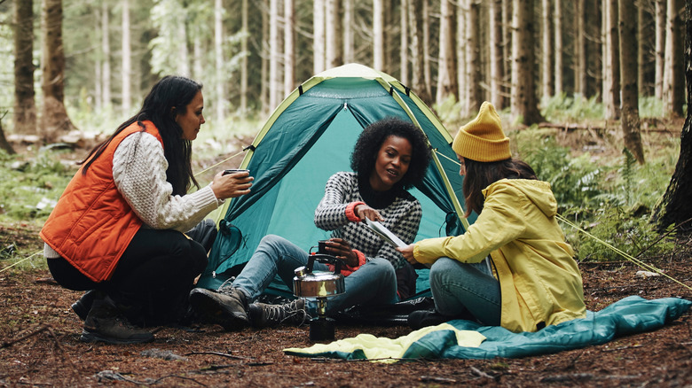 Group of diverse young female friends sitting at their forest campsite drinking coffee and reading a trail map