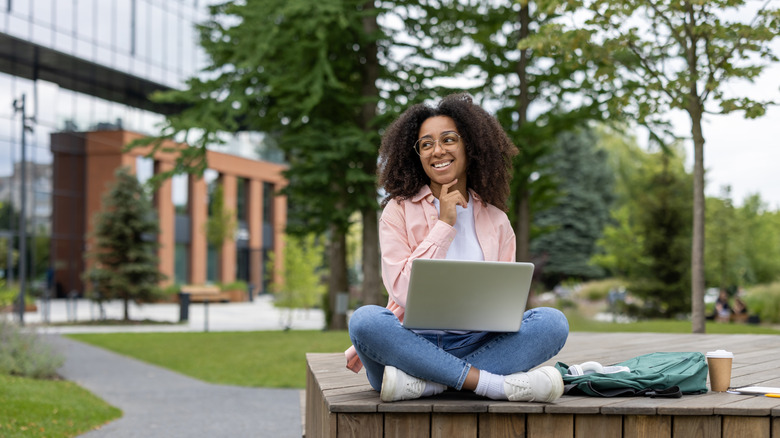 Happy female student sitting on campus bench with laptop