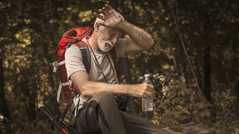 An older man wearing a backpack sitting in a forest with a bottle of water in hand