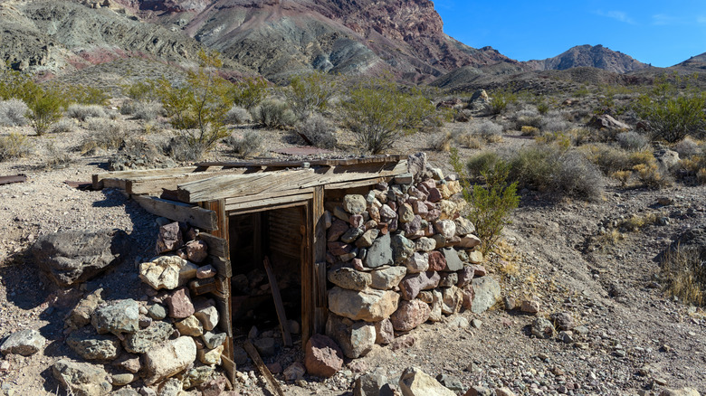 A mineshaft opening surrounded by rocks and wood and a rocky valley behind it with some mountains