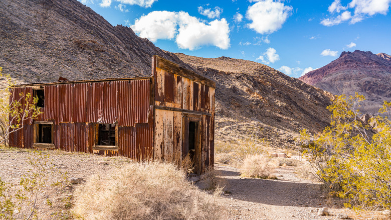 A rusty shack with a trail in front of it and mountains behind it