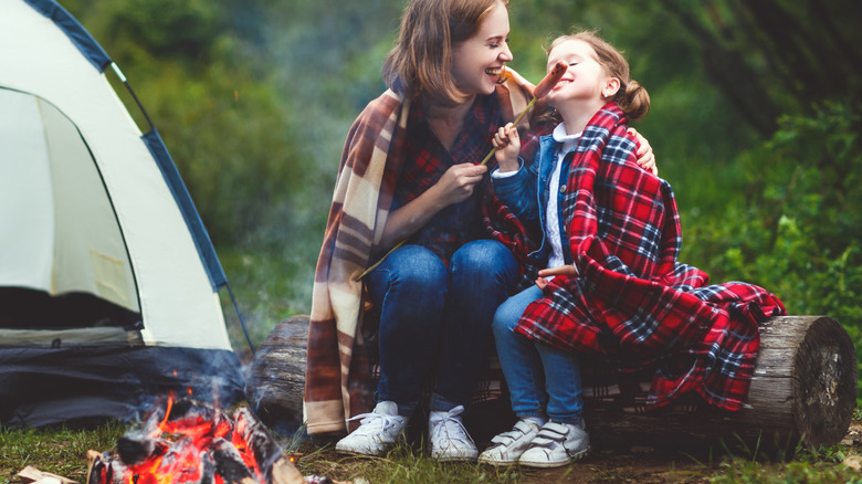 A mother and daughter eat a hotdog over a campfire.