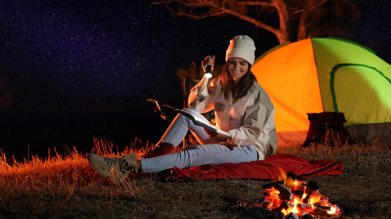 A woman near a tent holds up a flashlight so she can read.