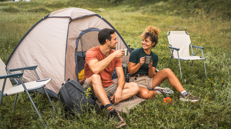 A couple sitting on the ground drinking coffee in front of a tent.