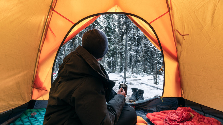 A man sitting in a sleeping bag inside of a tent on a cold snowy day.