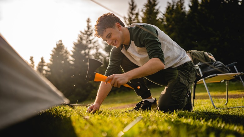 A young man hammers a tent peg into the ground with the back of a hatchet