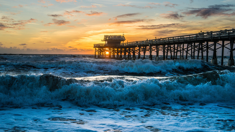 Aerial view of a beach with strong waves and the sun setting behind a pier