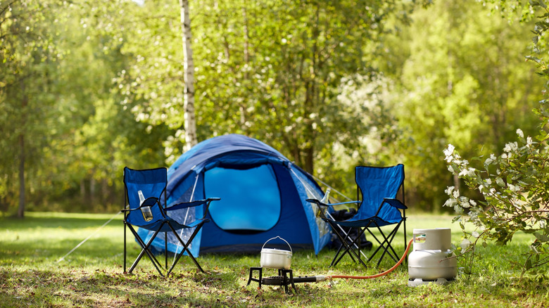 A blue tent and camping chairs in the woods next to a cooking setup.