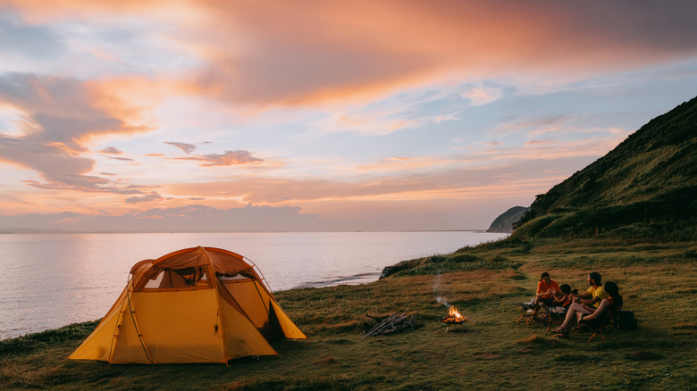 Campers beside a campfire and tent next to a lake at dusk.