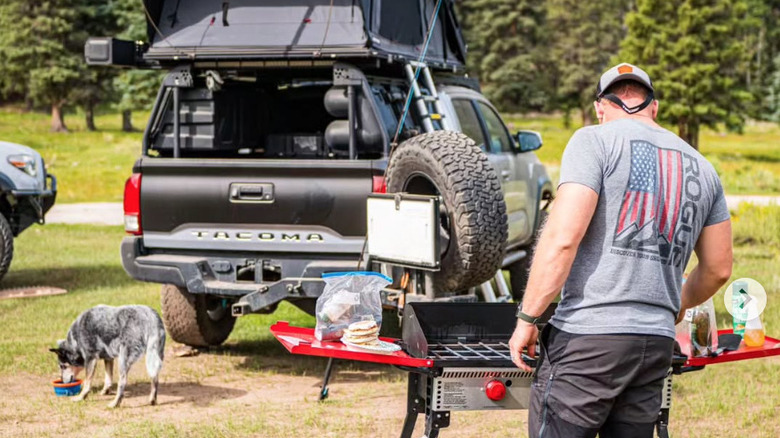 A man using his Camp Chef burner stove with a truck and dog in the background