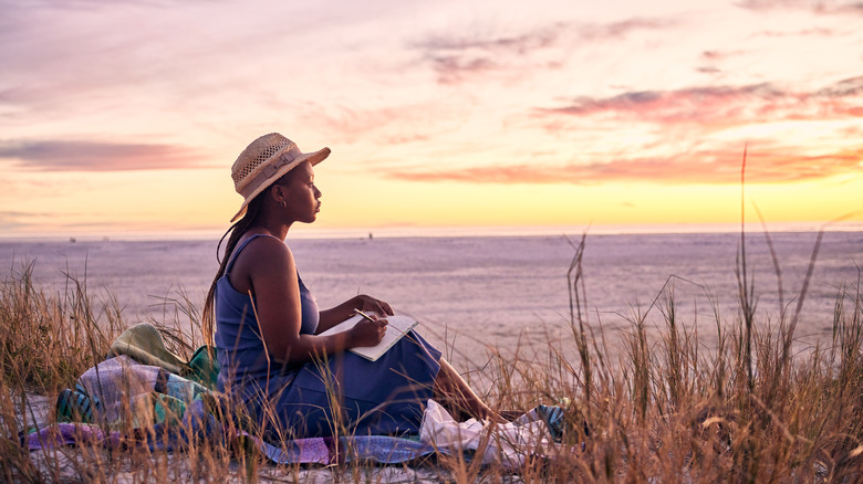 A woman serenely writes in a journal while the sun sets on the beach