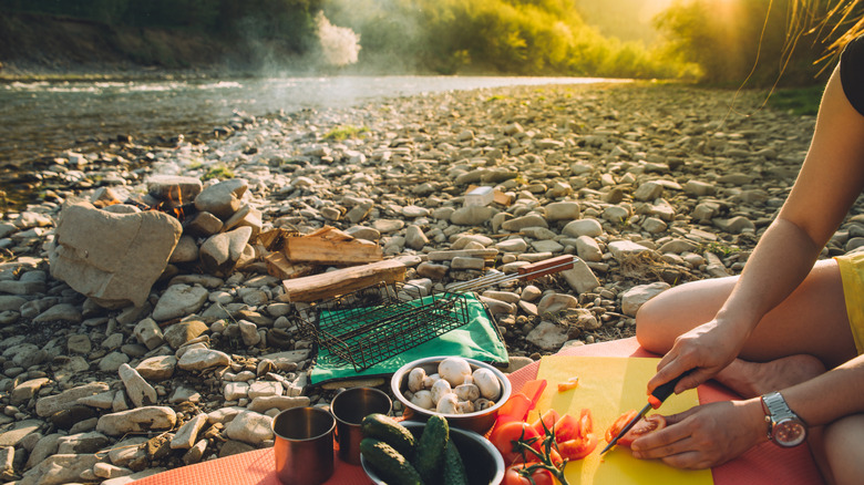 A woman's hands are shown cutting up vegetables while sitting along a rocky riverbed
