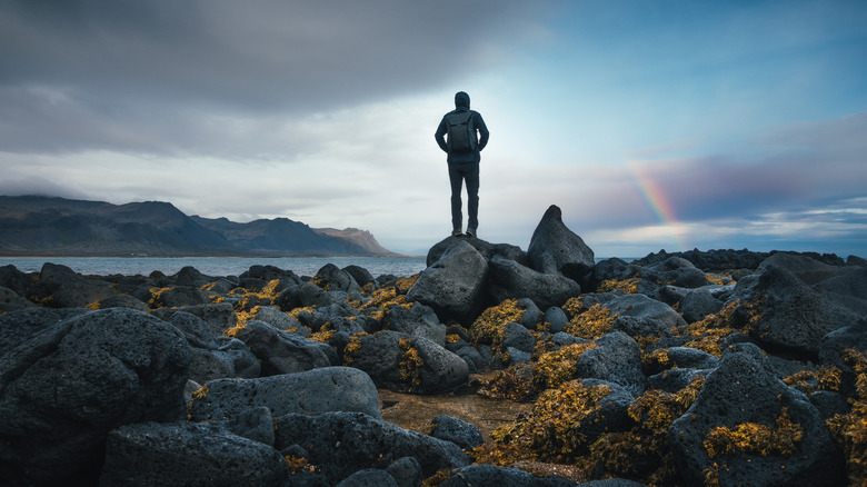 Backpacker stands on rocky shore.