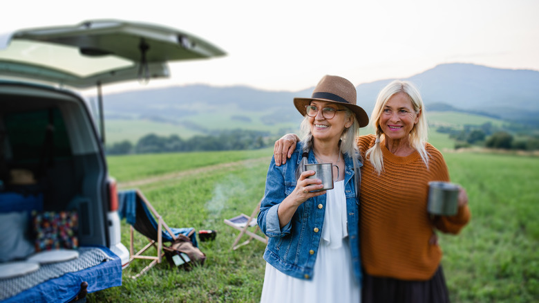 Two women drink from mugs in front of a campervan