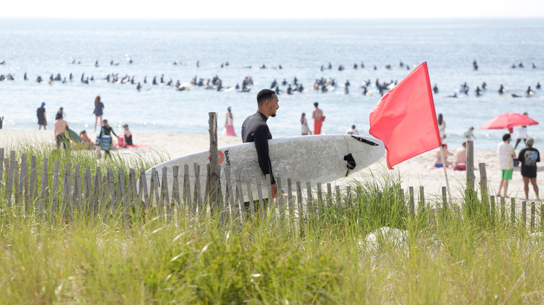 A surfer walking to the shore at Rockaway Beach near a red flag