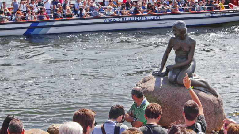 Crowds gather around the little mermaid statue in copenhagen as a boat sails past.