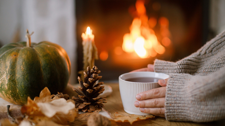 Hands holding cup of tea; pumpkin and autumn leaves on table on background of fireplace.