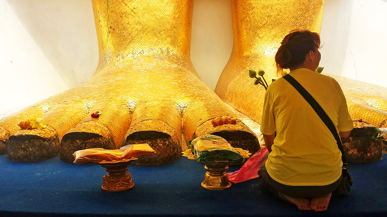 A Thai woman kneeling in front of the feet of a golden Buddha statue in a temple.
