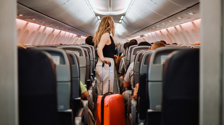 A passenger standing on a plane with a suitcase while other travelers are sitting down