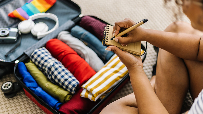 A person begins a list in a notebook near a packed suitcase