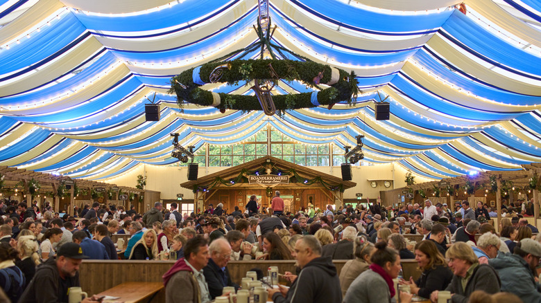 A beer hall with a blue and white tent ceiling filled with people eating and drinking