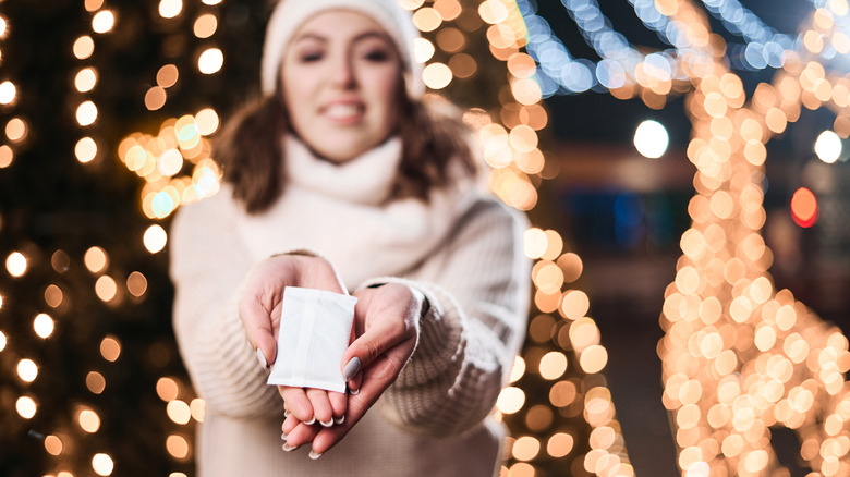 A woman in a sweater and cap holds out a hand warmer.