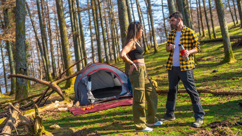 A man and woman converse near a tent in the woods