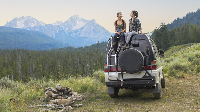 Two women sit on the roof an RV in front of rocky mountains