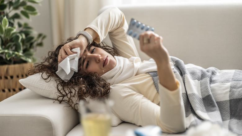 A sick woman laying on the couch holding medicine up and covering head with a tissue