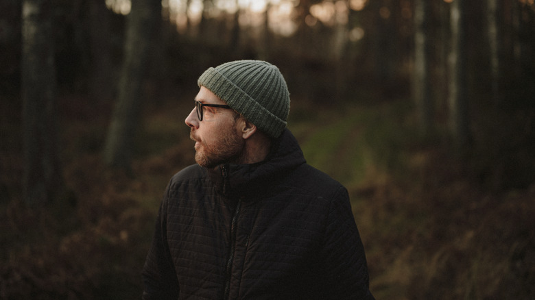 A bespectacled man in a beanie is alone in darkening woods
