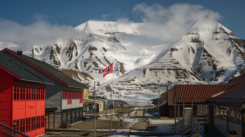 A small town with colorful wooden buildings, a raised Norwegian flag, and snow covered mountains in the background