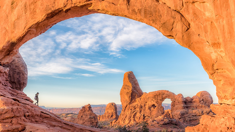 hiker under massive natural stone arch in view of another arch at Arches National Park in Utah