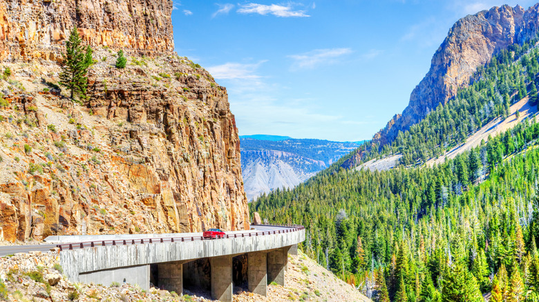 car driving on a road curving around a beige cliff, evergreens on an adjacent rocky slope