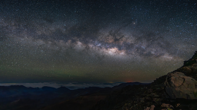 The Milky Way stretches brightly across the night sky above Haleakalā, illuminating rugged volcanic cliffs and a faint sea of clouds below