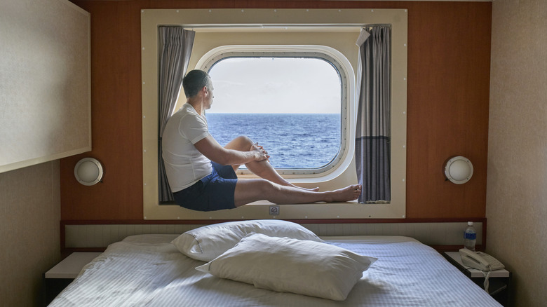 A man gazes out a porthole in his cruise ship cabin
