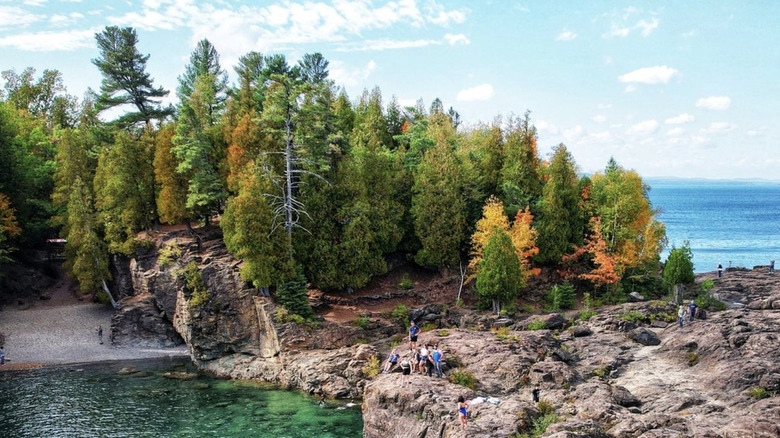 People explore a rocky shoreline with evergreens and autumnal foliage on the turquoise waters of Lake Superior, Isle Royale National Park