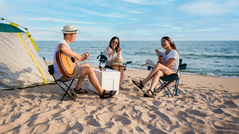 Group of happy Asian friends playing guitar and singing with clap while picnic and camping on the beach in outdoors